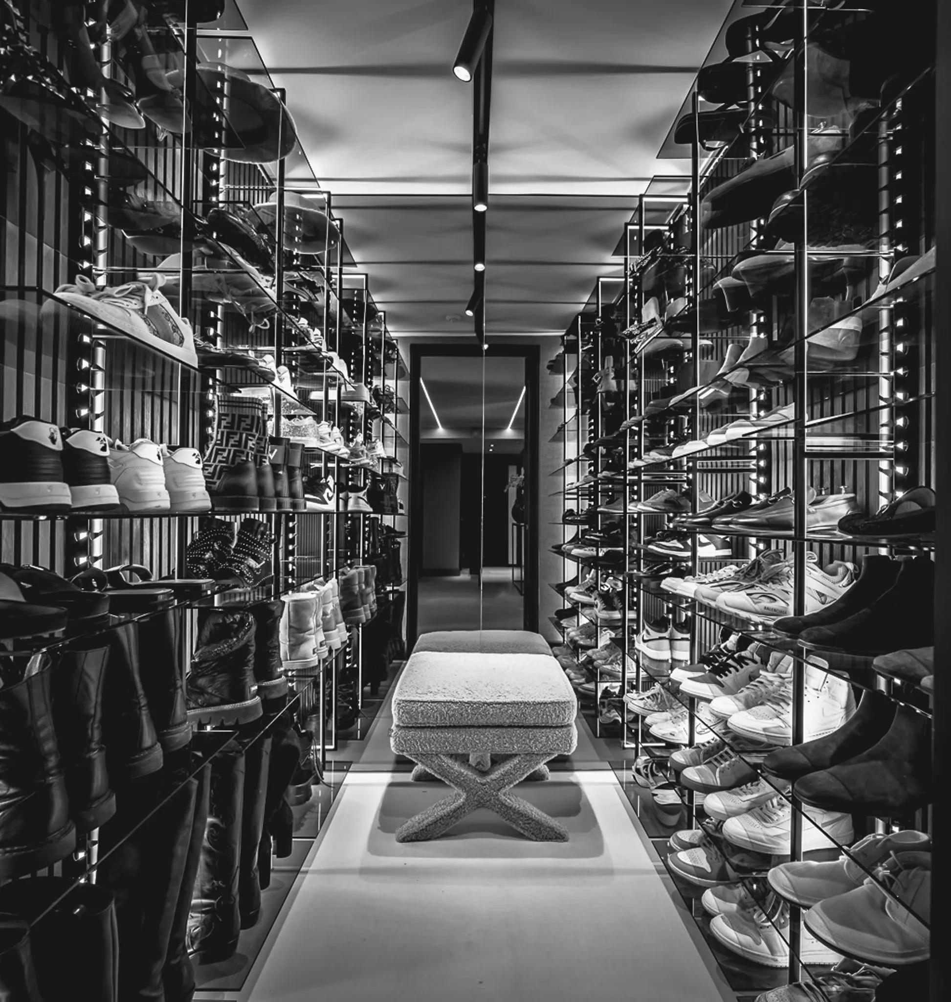 Inloopkast met spiegelwanden en verlichte glazen schappen vol schoenen aan weerszijden van een smalle gang, in zwart-wit